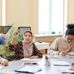Muslim woman attending language lessons for immigrants raising hand to ask teacher question about task
